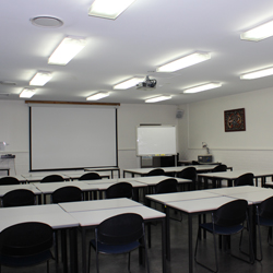 There are rows of desks set up facing the front of the room. The front of the room has a whiteboard and a projector set up.