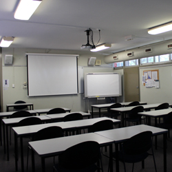 There are four rows of desks set up facing the front of the room. The front of the room has a whiteboard and a projector set up.