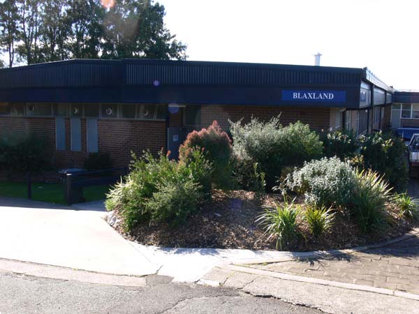 Single storey brick accommodation block external view. Circle garden bed with plants and building behind it. Blaxland sign located on roof.