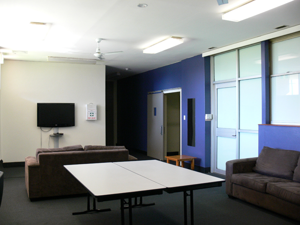 Room with two 2-seater lounges. Blue feature wall to right of room and two doors. TV mounted on wall to left of room. Tables in middle of room.