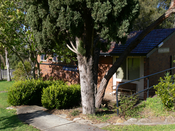 Small single storey brick accommodation cottage. Large tree surrounded by bushes with building behind it. Evans cottage sign mounted on wall of building.