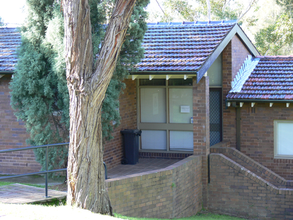 Small single storey brick accommodation cottage with large tree at front. There are two staircase entrances to the front door.