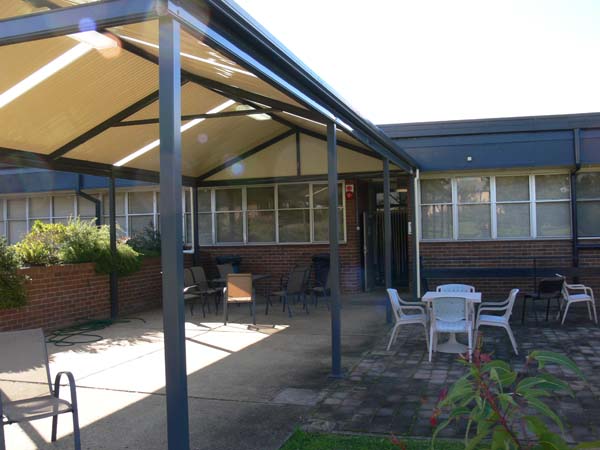 Outdoor seating area outside entry to large brick accommodation building. It is under an awning and several tables and chairs. Entrance at the end of awning.