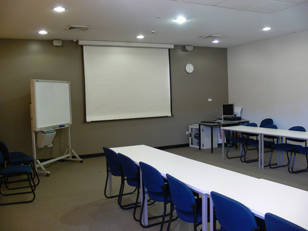 Training room set up with two rows of tables and chairs. Projector screen on back wall with whiteboard to the left.