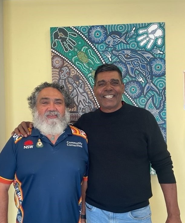 Two men standing, arms on each other's shoulder in front of an indigenous artwork