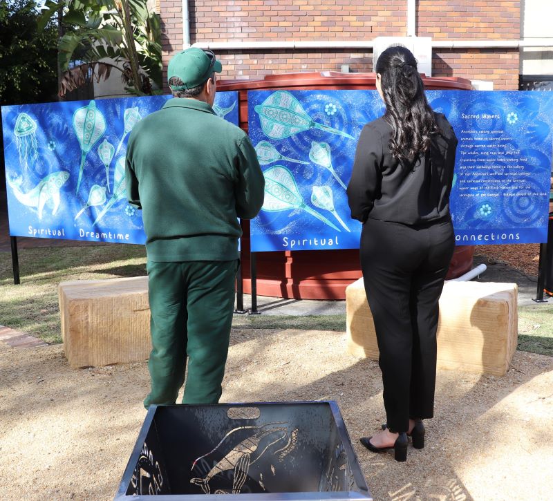 Foreground: inmate in green tshirt and shorts stands next to uniformed custodial officer, Aboriginal artwork in the background  