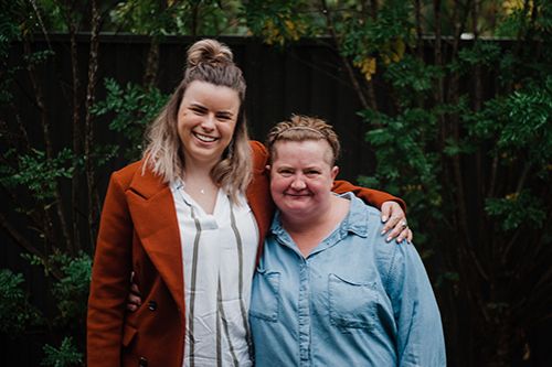 Two females standing next to each other smiling with an arm over each others shoulder