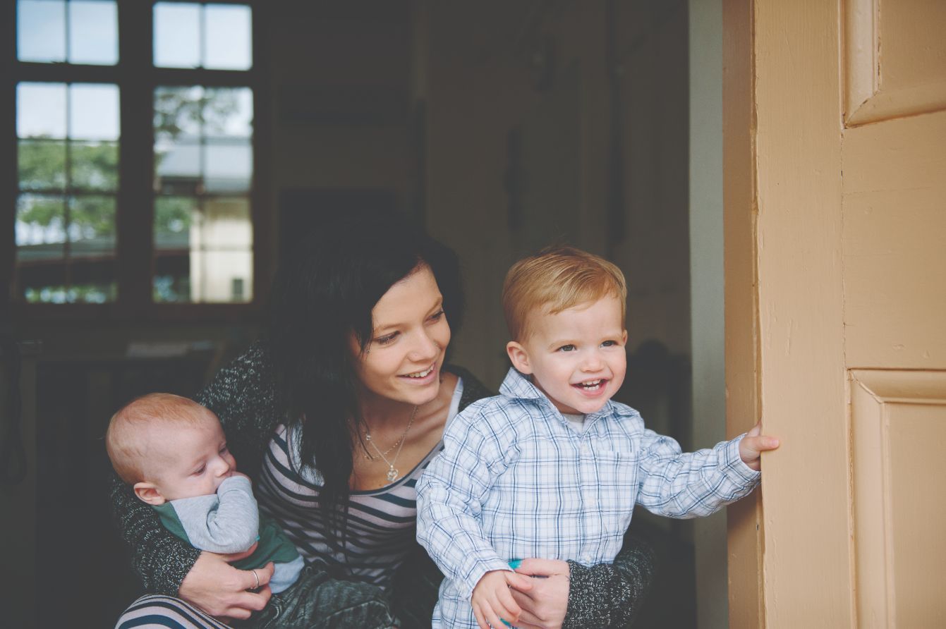 A female adult holding and infant child and smiling crouching down to speak with her toddler